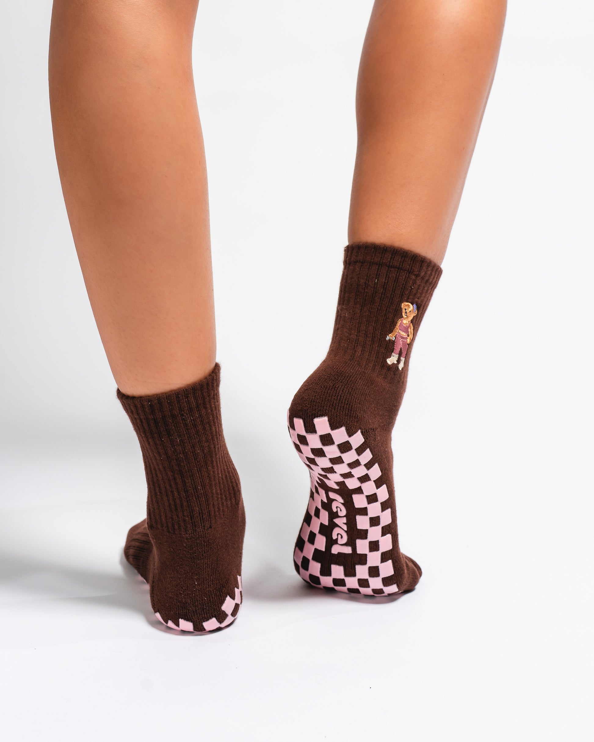 A person wearing Revel Athletic's The Teddy Grip Sock—a brown crew sock with a small teddy embroidery on the side and a pink grippy checker bottom—stands on a white background.