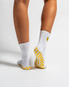 A person stands on tiptoe against a plain white background, wearing Revel Athletic’s The I’m Fine Grip Sock—white crew Pilates socks featuring yellow checkered soles and a yellow smiley face logo on the back.