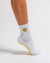 A person wearing The I'm Fine Grip Sock by Revel Athletic—featuring a white base, a yellow smiley face design, and yellow grips on the bottom—stands on one foot against a white background.