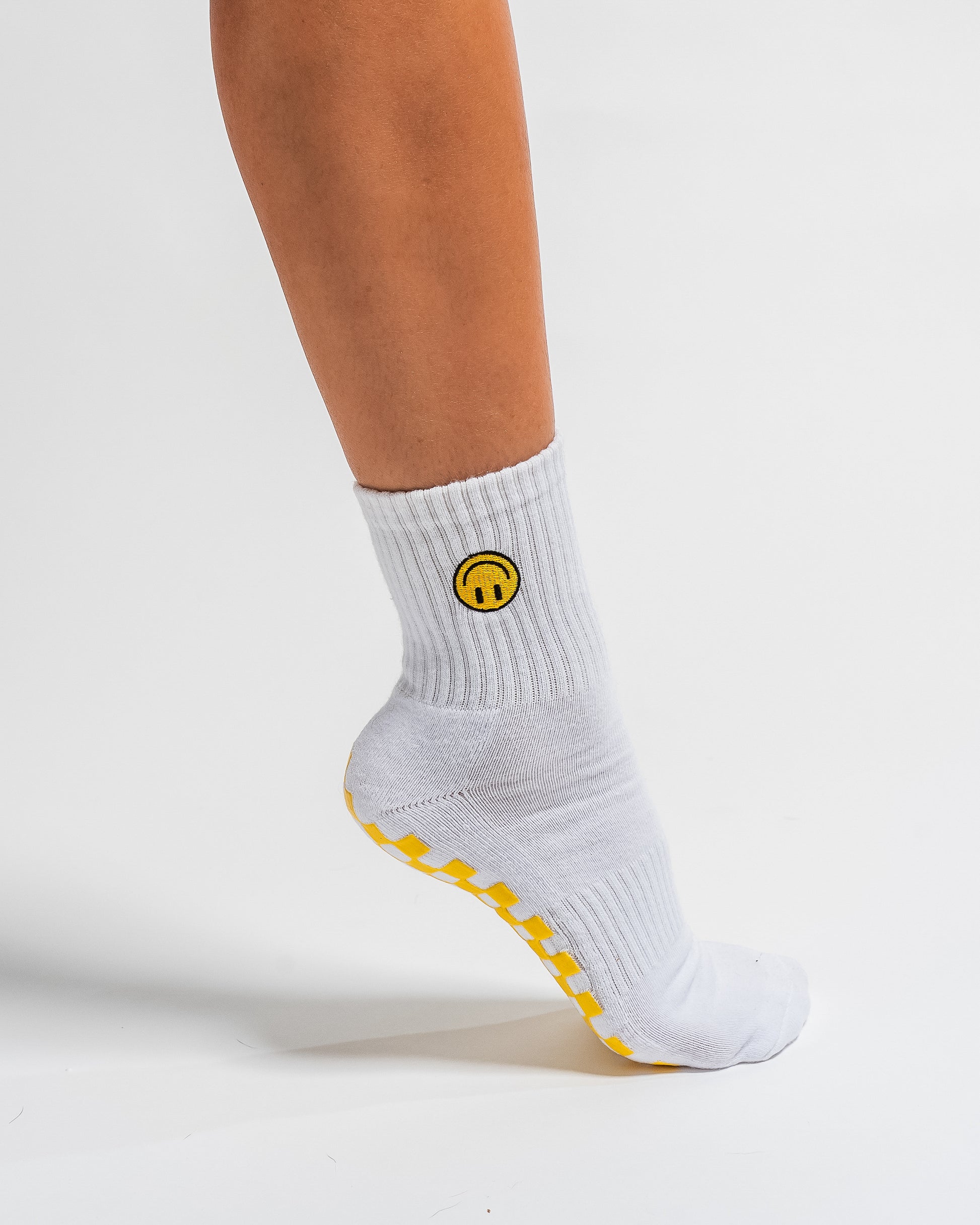 A person wearing The I'm Fine Grip Sock by Revel Athletic—featuring a white base, a yellow smiley face design, and yellow grips on the bottom—stands on one foot against a white background.