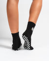 A person wearing Revel Athletic’s The All Nighter Grip Socks, featuring a black design with white checkerboard-patterned soles, stands on a white surface with heels slightly raised—perfect for Pilates or yoga practice.