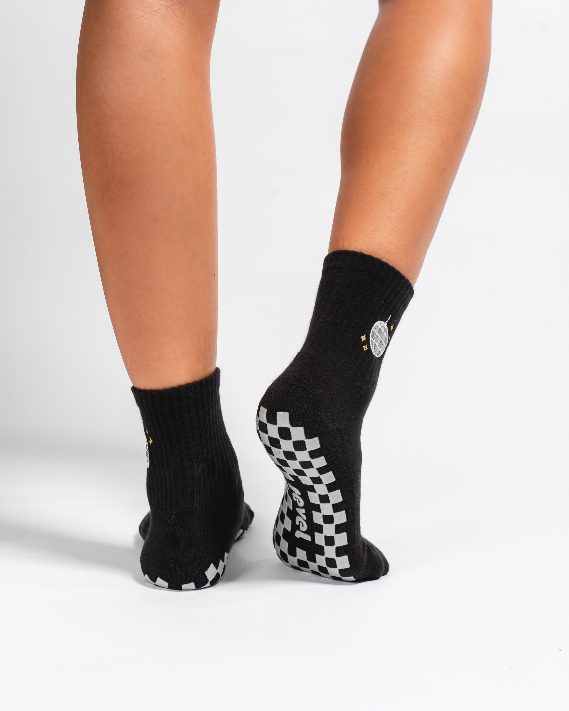 A person wearing Revel Athletic’s The All Nighter Grip Socks, featuring a black design with white checkerboard-patterned soles, stands on a white surface with heels slightly raised—perfect for Pilates or yoga practice.