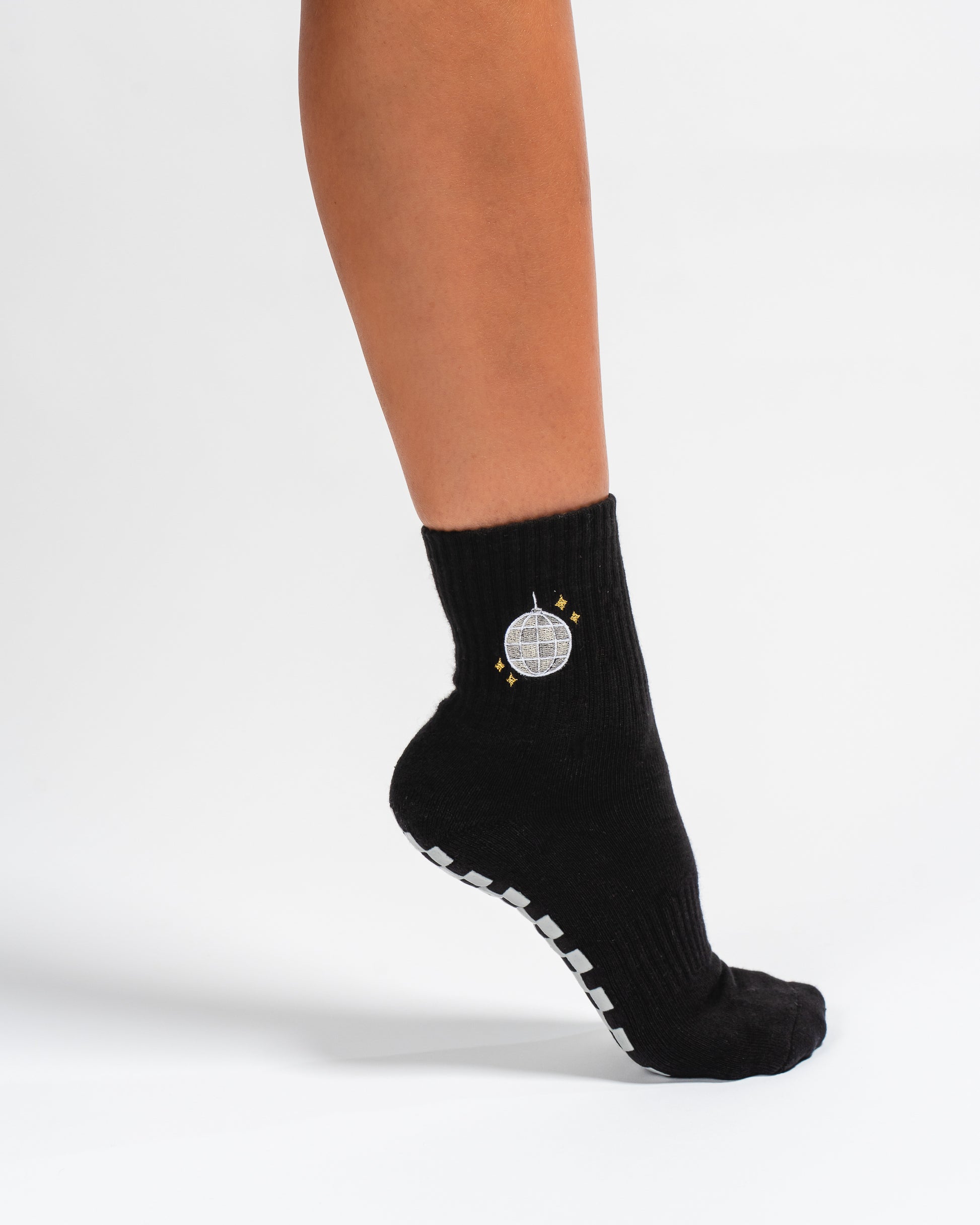A person wearing Revel Athletic's The All Nighter Grip Sock, featuring a black base with silver disco ball and gold star designs, stands on their toes. The sock’s white grip strips on the sole provide stability, making it ideal for yoga or studio workouts.