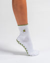 A person wearing The Extra Dirty Grip Sock by Revel Athletic—a white sock featuring a green olive embroidery on the side and a green scalloped edge—stands on tiptoe. Designed for Barre or Pilates, this sock offers green grip details on the sole to deliver extra stability during your workout.