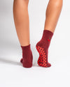 A person is shown from the calf down against a white background, wearing Revel Athletic’s The Very Cherry Grip Sock. These socks feature vibrant cherry embroidery stitched on the sides, red checkerboard pattern non-slip grips on the soles for ideal yoga traction, and "Lucky" printed near the toes.