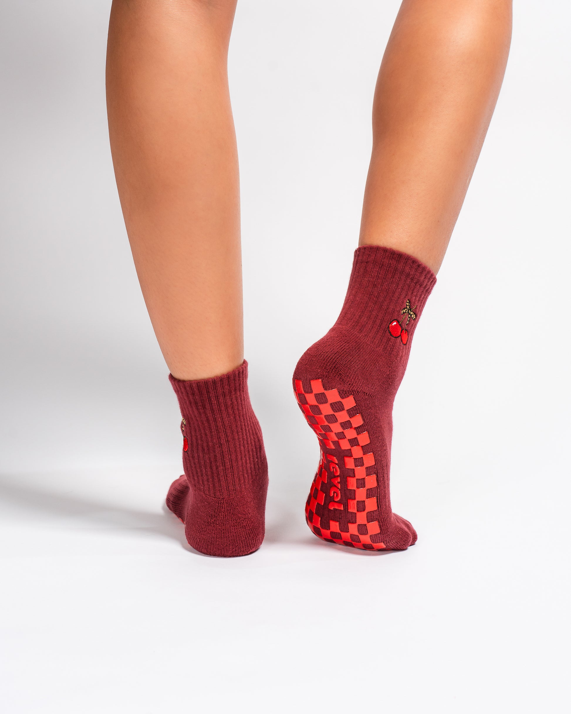 A person is shown from the calf down against a white background, wearing Revel Athletic’s The Very Cherry Grip Sock. These socks feature vibrant cherry embroidery stitched on the sides, red checkerboard pattern non-slip grips on the soles for ideal yoga traction, and "Lucky" printed near the toes.