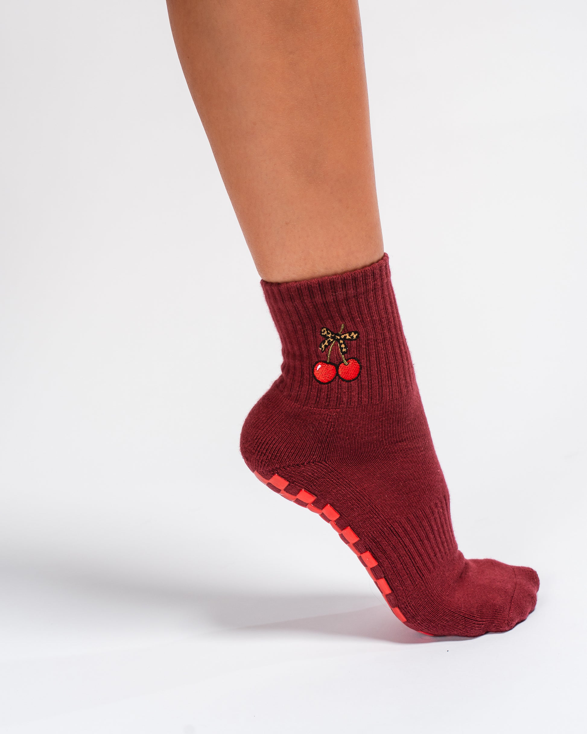 A person wearing The Very Cherry Grip Sock by Revel Athletic—featuring maroon fabric with cherry embroidery and red grips on the sole, ideal for yoga—stands with their foot slightly lifted against a white background.