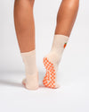 A person wearing The Just Peachy Grip Sock by Revel Athletic, featuring cream-colored fabric with orange checkerboard patterns and small strawberry designs, stands on a white background. These grip socks blend style and functionality, making them perfect for your workout routine.