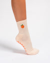 A person wearing The Just Peachy Grip Sock by Revel Athletic—a light beige pilates sock featuring a peach fruit design on the side and textured peach-colored grips on the sole—stands on tiptoe against a white background.