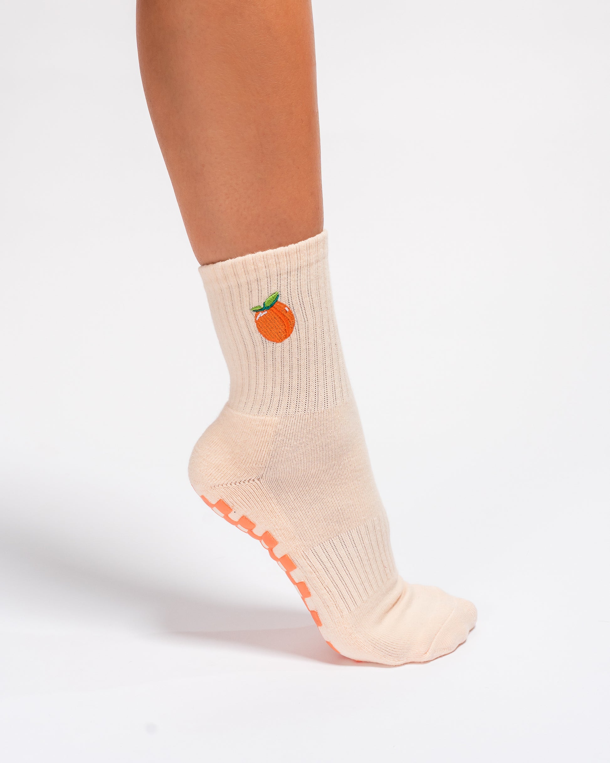 A person wearing The Just Peachy Grip Sock by Revel Athletic—a light beige pilates sock featuring a peach fruit design on the side and textured peach-colored grips on the sole—stands on tiptoe against a white background.