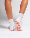 Two legs are wearing "The Spritz Grip Sock" by Revel Athletic—white crew Pilates grip socks featuring orange checkered grip patterns on the soles. A small orange flower design decorates the back of one sock, adding a playful touch. The feet are positioned on a white background.