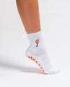 A person standing on tiptoe against a white background wears The Spritz Grip Sock by Revel Athletic, featuring a white design with an embroidered cocktail glass and bright orange grip dots on the sole.