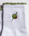 The Extra Dirty Grip Sock by Revel Athletic features white ribbed fabric with a green scalloped edge, an embroidered green olive with a red pimento and cocktail stick—perfect for bringing a playful touch to your Barre, Pilates, or Yoga practice.