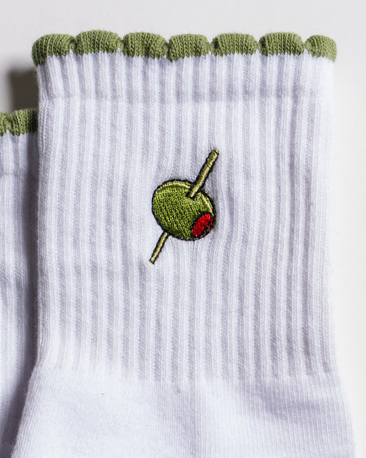 The Extra Dirty Grip Sock by Revel Athletic features white ribbed fabric with a green scalloped edge, an embroidered green olive with a red pimento and cocktail stick—perfect for bringing a playful touch to your Barre, Pilates, or Yoga practice.