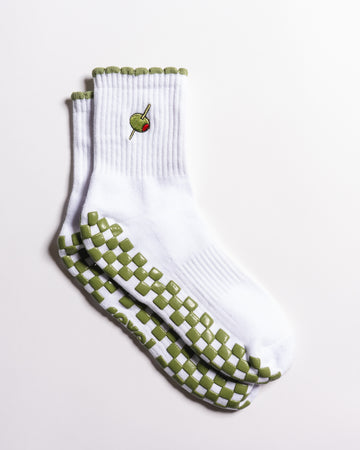 Introducing The Extra Dirty Grip Sock by Revel Athletic: a white crew sock featuring green and white checkerboard grips on the soles for superior traction during Barre, Pilates, or Yoga, and accented with a small embroidered olive (complete with toothpick) near the top.