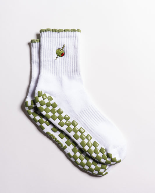 Introducing The Extra Dirty Grip Sock by Revel Athletic: a white crew sock featuring green and white checkerboard grips on the soles for superior traction during Barre, Pilates, or Yoga, and accented with a small embroidered olive (complete with toothpick) near the top.