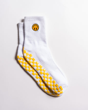 The I'm Fine Grip Sock by Revel Athletic is a pair of white socks featuring a yellow smiley face near the top and a yellow checkerboard pattern on the soles, designed for extra traction and perfect as Pilates socks; they're displayed on a white surface.