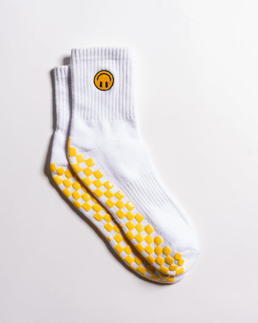 The I'm Fine Grip Sock by Revel Athletic is a pair of white socks featuring a yellow smiley face near the top and a yellow checkerboard pattern on the soles, designed for extra traction and perfect as Pilates socks; they're displayed on a white surface.