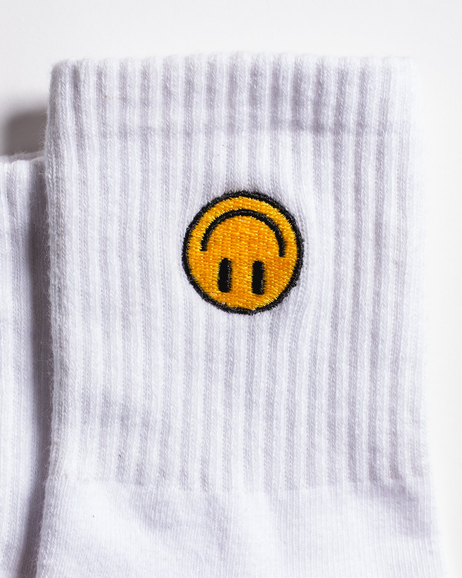 The I'm Fine Grip Sock by Revel Athletic features a white ribbed design with an embroidered yellow smiley face—sporting an upside-down mouth and outlined in black—on the upper part of the sock.