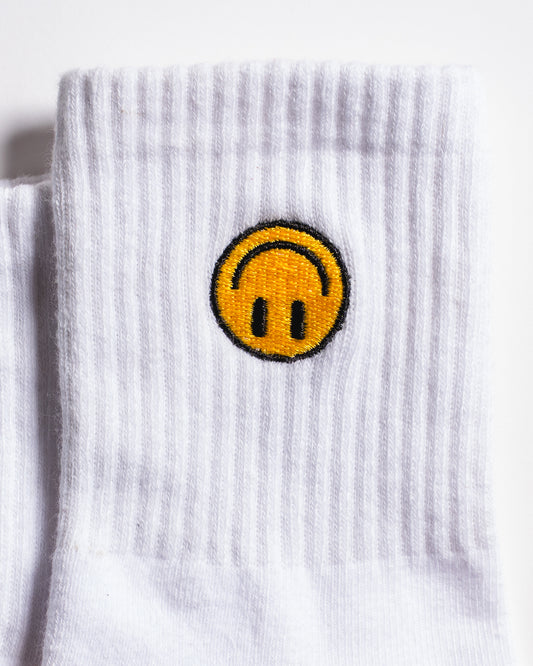 The I'm Fine Grip Sock by Revel Athletic features a white ribbed design with an embroidered yellow smiley face—sporting an upside-down mouth and outlined in black—on the upper part of the sock.
