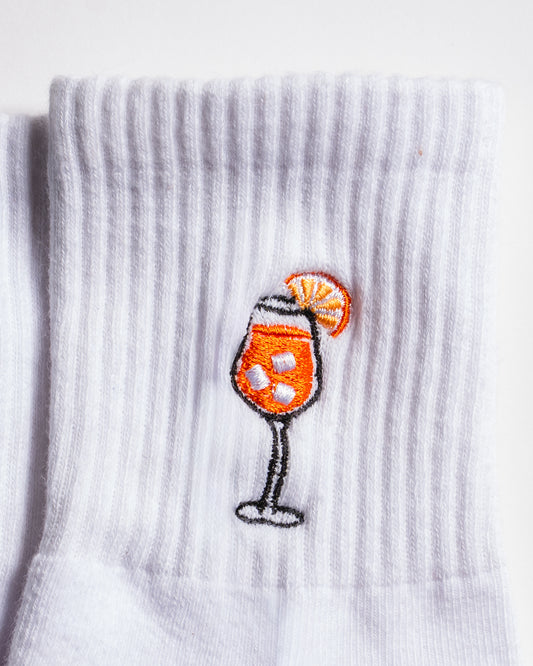 A detailed look at The Spritz Grip Sock by Revel Athletic—a white ribbed sock adorned with an embroidered Aperol Spritz in a tall glass, complete with ice cubes and an orange slice garnish—makes a playful addition to your Pilates grip socks collection.