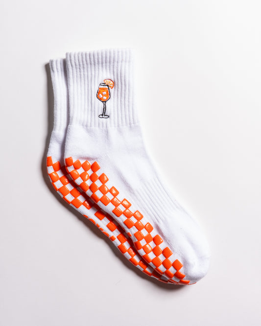 Introducing The Spritz Grip Sock by Revel Athletic: a pair of white grip socks featuring an orange checkerboard pattern on the soles for enhanced traction—ideal for Pilates or barre classes. Finished with a small embroidered orange cocktail glass and slice of orange near the top, these playful socks bring Aperol Spritz-inspired flair to your workout.