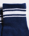 Close-up of the top part of The Country Club Grip Sock by Revel Athletic in navy blue, featuring two white horizontal stripes bordered by thin navy lines near the cuff. The socks are laid flat against a light background.
