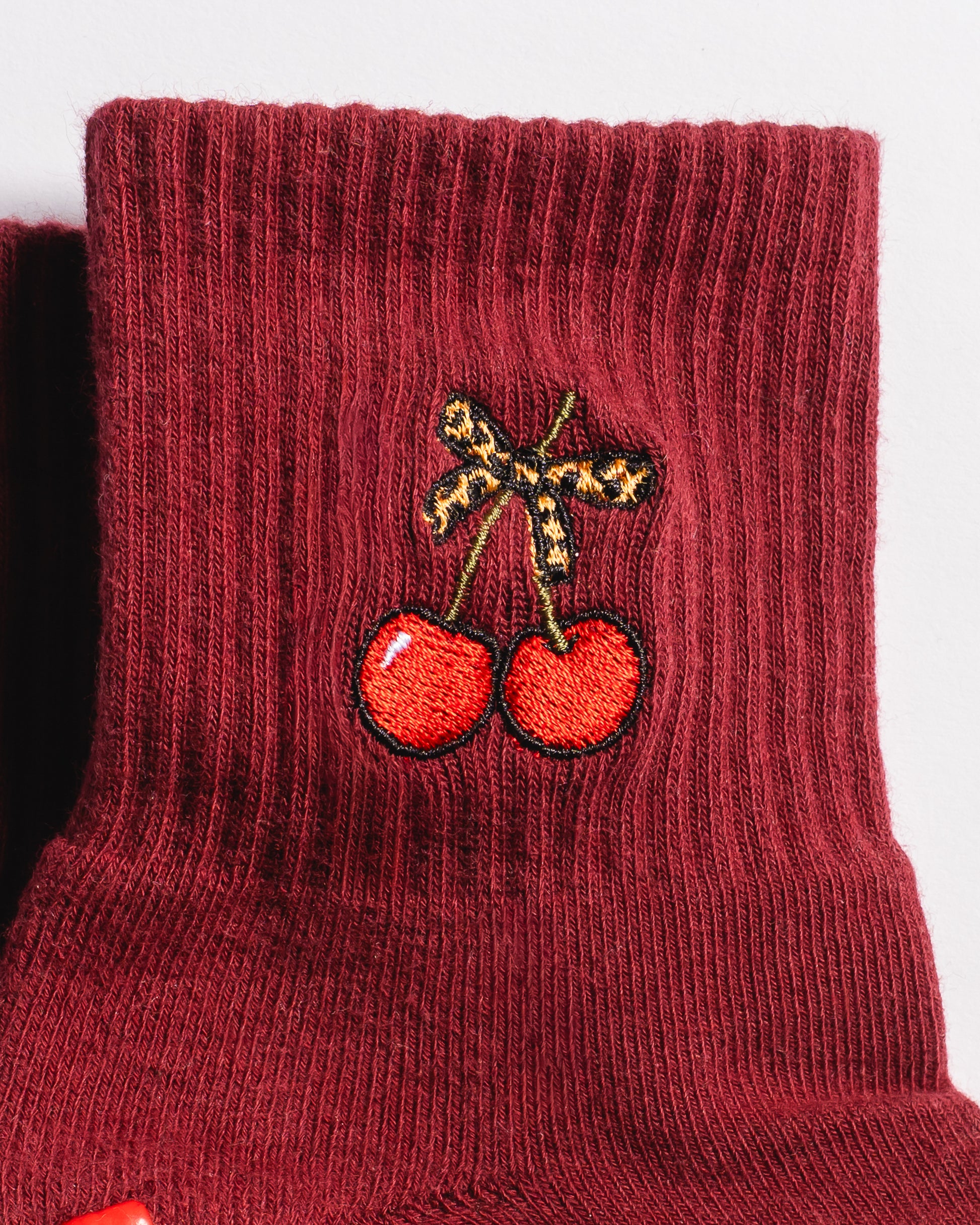 A close-up of The Very Cherry Grip Sock by Revel Athletic showcases a maroon, ribbed athletic sock featuring an embroidered design of two red cherries with a brown bow near the top. These cherry-themed grip socks offer playful style and comfort—perfect for adding a cheerful touch to your Pilates or yoga collection.