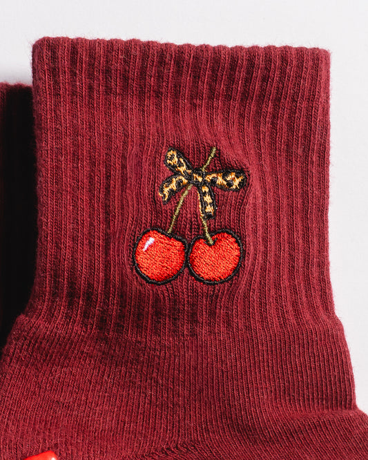 A close-up of The Very Cherry Grip Sock by Revel Athletic showcases a maroon, ribbed athletic sock featuring an embroidered design of two red cherries with a brown bow near the top. These cherry-themed grip socks offer playful style and comfort—perfect for adding a cheerful touch to your Pilates or yoga collection.