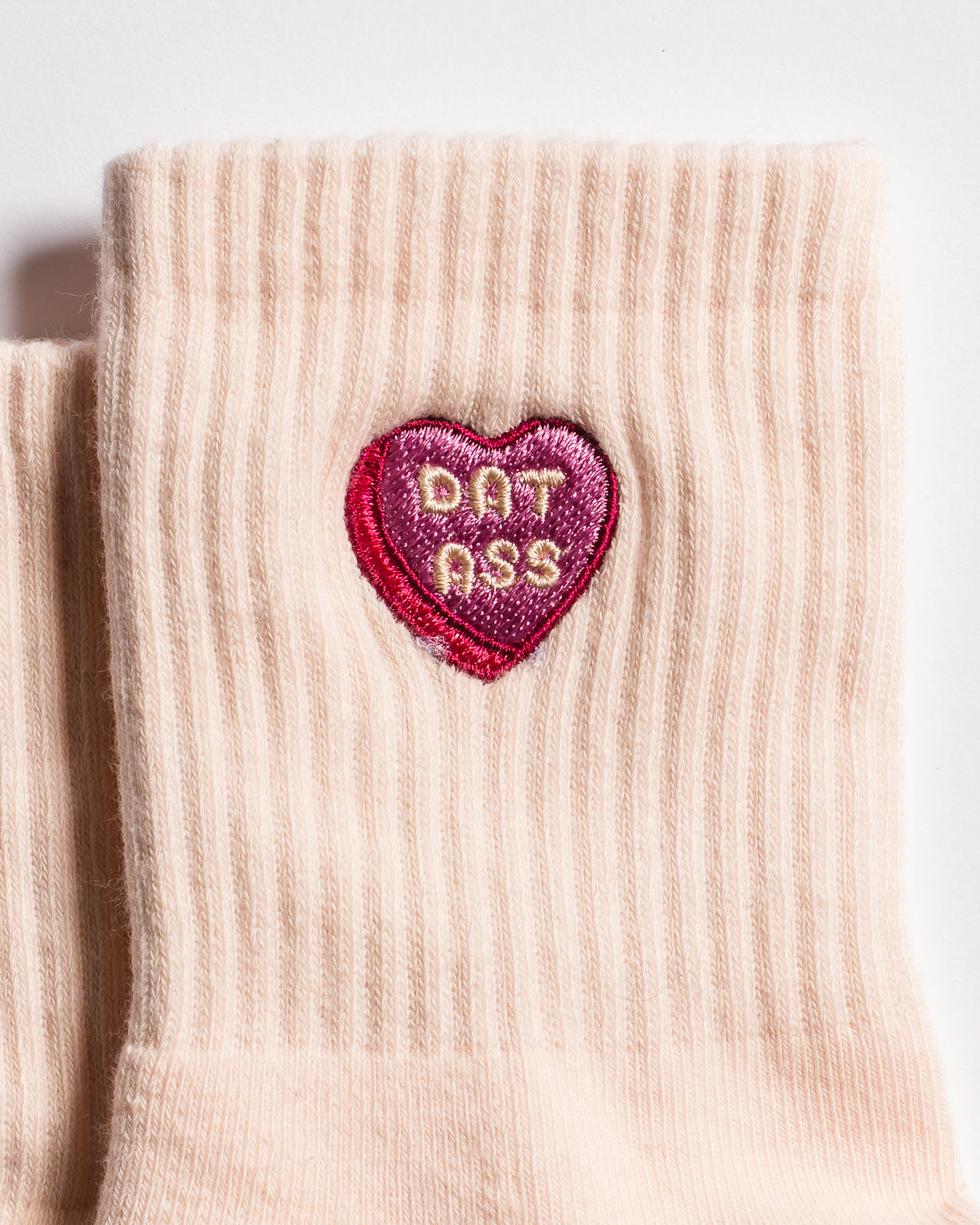 A close-up of The Sweet Nothings Grip Sock by Revel Athletic, featuring a beige design with a pink and red heart-shaped patch embroidered with "DAT ASS" in the center.
