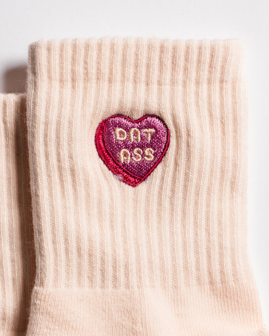 A close-up of The Sweet Nothings Grip Sock by Revel Athletic, featuring a beige design with a pink and red heart-shaped patch embroidered with "DAT ASS" in the center.