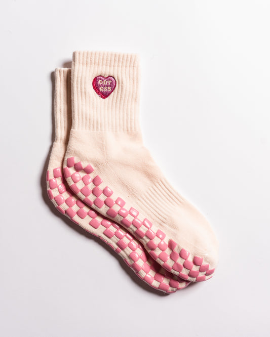 Introducing The Sweet Nothings Grip Sock by Revel Athletic—a pair of light pink grippy socks featuring pink-and-white checkerboard non-slip soles and a pink heart patch on the ankle that reads "BAD ASS." Shown against a white background, these socks are perfect for Pilates, barre, or your next workout.