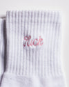 A close-up of The Pardon My French Grip Sock (Petal Pink) by Revel Athletic, a white ribbed Pilates grip sock featuring "luck" embroidered in pink near the top cuff, designed for both comfort and stability with added style during your workout.