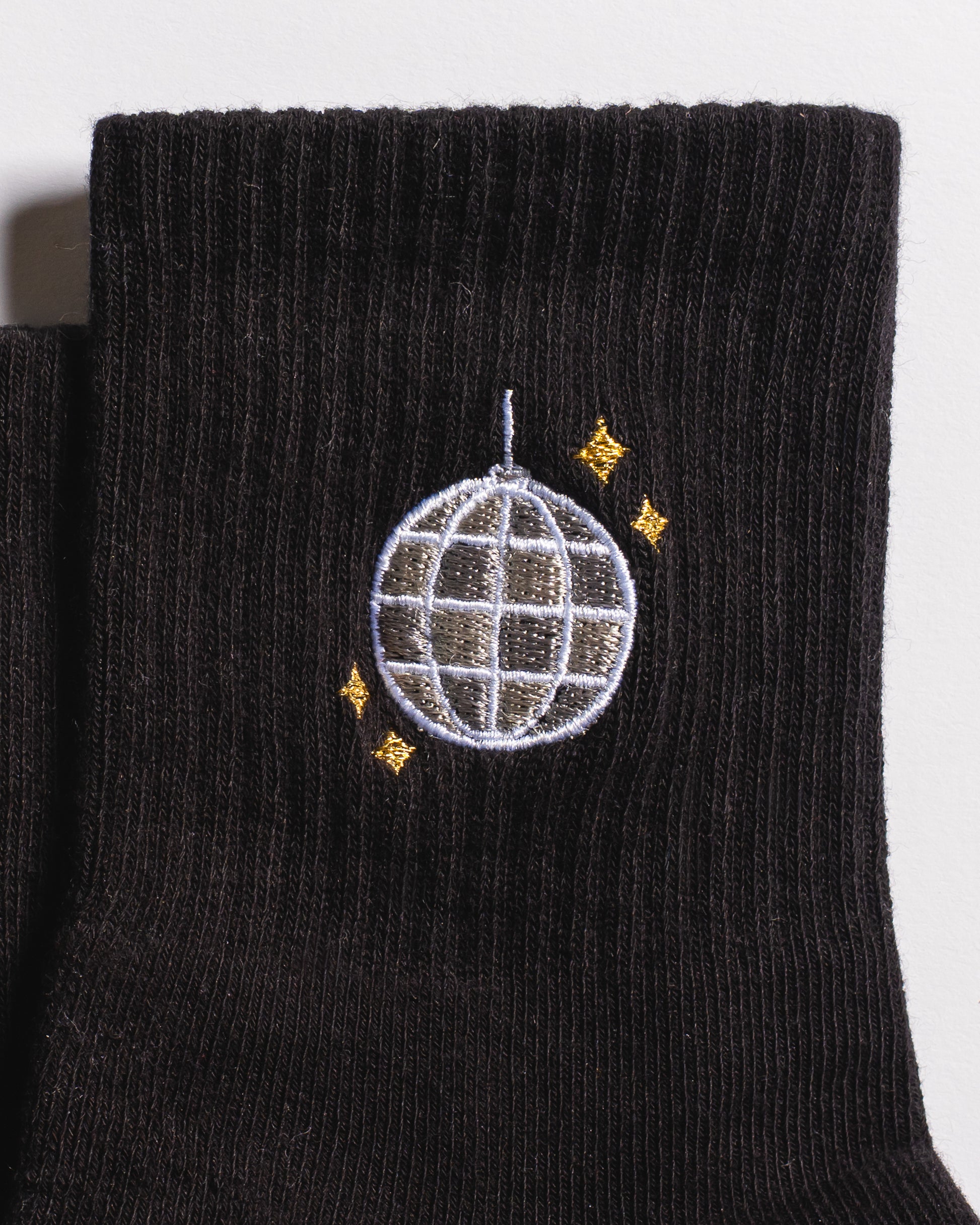 A close-up of The All Nighter Grip Sock by Revel Athletic—a black grip sock ideal for yoga or Pilates, adorned with an embroidered silver disco ball and four small yellow stars.