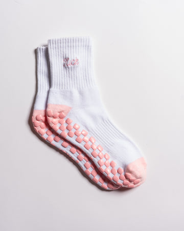 A pair of Revel Athletic’s The Pardon My French Grip Socks (Petal Pink) in white, featuring petal pink non-slip grips on the soles and toes, with “meh” embroidered near the top in pink. The socks are neatly stacked on a plain white background.