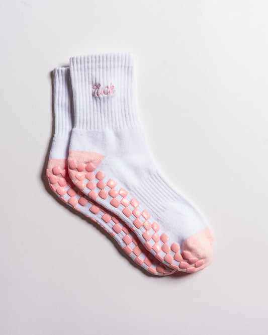 A pair of Revel Athletic’s The Pardon My French Grip Socks (Petal Pink) in white, featuring petal pink non-slip grips on the soles and toes, with “meh” embroidered near the top in pink. The socks are neatly stacked on a plain white background.