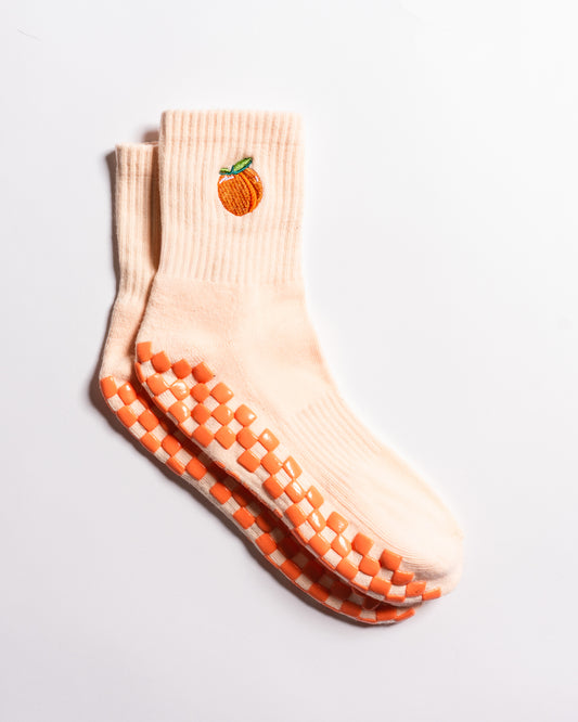 Introducing The Just Peachy Grip Sock by Revel Athletic: a pair of light peach socks featuring vibrant orange non-slip grips on the soles and an embroidered peach fruit detail near the top. Ideal for pilates or daily wear, these grip socks provide comfort and style, shown here against a white background.
