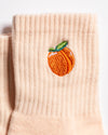 The Just Peachy Grip Sock by Revel Athletic is a beige ribbed sock featuring an embroidered orange peach with a green leaf near the top. These pilates socks are displayed lying flat on a white background.
