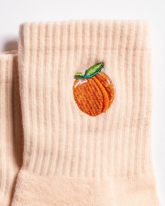 The Just Peachy Grip Sock by Revel Athletic is a beige ribbed sock featuring an embroidered orange peach with a green leaf near the top. These pilates socks are displayed lying flat on a white background.