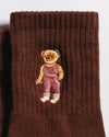 Introducing The Teddy Grip Sock by Revel Athletic: Crafted from brown knit fabric, this cozy sock features a charming embroidered teddy bear dressed in pink overalls and a blue hat stitched on the surface. The grippy checker bottom is ideal for Pilates or relaxing at home, while the playful teddy embroidery adds a delightful touch.