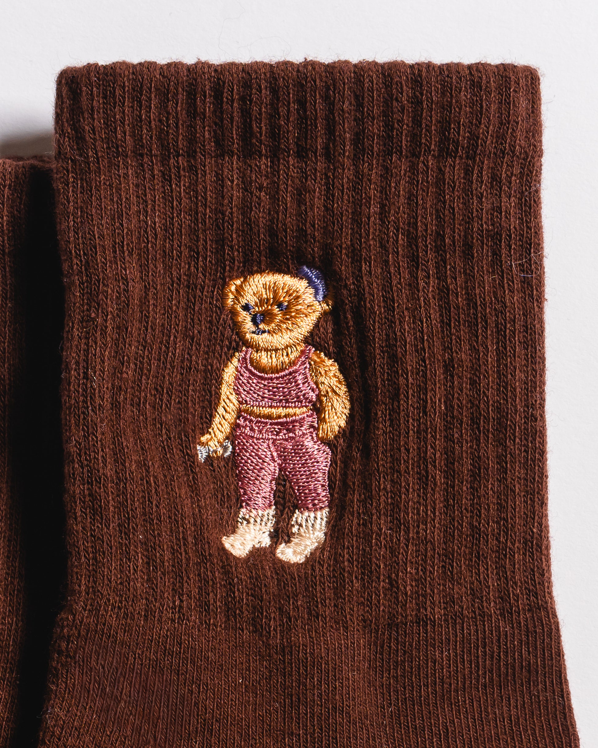 Introducing The Teddy Grip Sock by Revel Athletic: Crafted from brown knit fabric, this cozy sock features a charming embroidered teddy bear dressed in pink overalls and a blue hat stitched on the surface. The grippy checker bottom is ideal for Pilates or relaxing at home, while the playful teddy embroidery adds a delightful touch.
