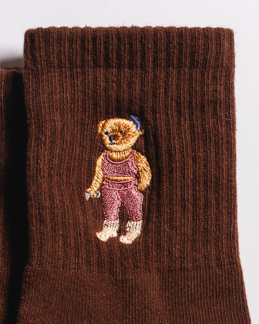 Introducing The Teddy Grip Sock by Revel Athletic: Crafted from brown knit fabric, this cozy sock features a charming embroidered teddy bear dressed in pink overalls and a blue hat stitched on the surface. The grippy checker bottom is ideal for Pilates or relaxing at home, while the playful teddy embroidery adds a delightful touch.