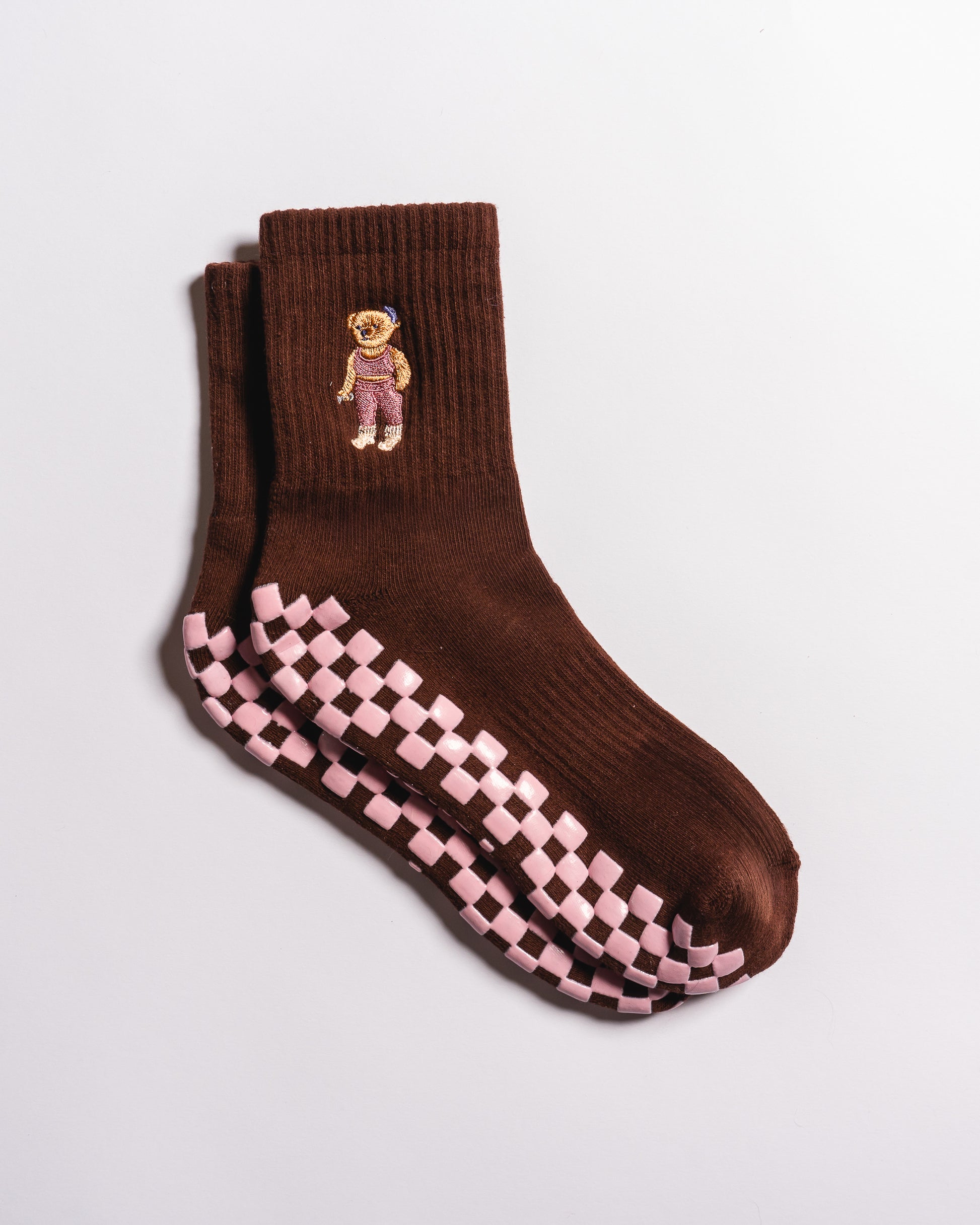 Introducing The Teddy Grip Sock by Revel Athletic: brown socks featuring a pink and white grippy checker-patterned sole and an embroidered teddy bear in a purple outfit near the cuff. These charming socks, displayed flat against a white background, are perfect for Pilates or lounging in style.