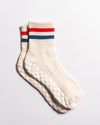 Two pairs of The Miss Americana Grip Socks by Revel Athletic, featuring a cream color with red and blue stripes near the top and white grip dots on the soles, are laid flat on a white background, slightly overlapping each other.