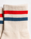 Close-up of Revel Athletic's "The Miss Americana Grip Sock" in cream, featuring ribbed cuffs and a horizontal red stripe above a blue stripe near the top. The non-slip socks are placed side by side on a white background.