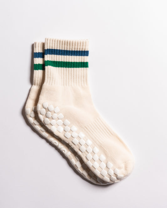 Two cream-colored "The Varsity Grip Sock" pairs from Revel, featuring green and blue stripes at the top, are stacked together. Their soles display white rubber grips in a grid pattern for enhanced stability during yoga or Pilates sessions. The grip socks are shown against a plain white background.