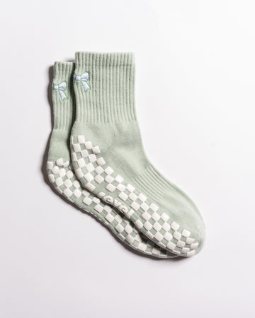 A pair of Revel Athletic "The Tied & True Grip Sock" in light green, featuring ribbed cuffs, small blue bows near the top, and white non-slip grip patterns on the soles.