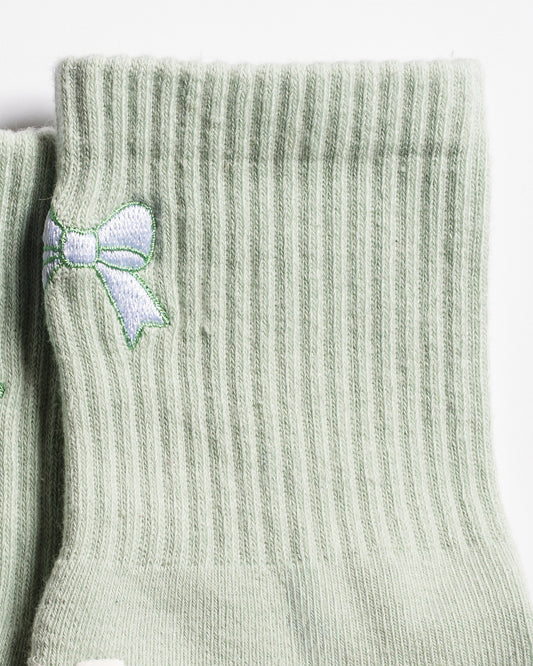 Close-up of The Tied & True Grip Socks by Revel Athletic in a light green ribbed design, featuring a small embroidered white and green bow near the top. The socks are positioned side by side on a white background.