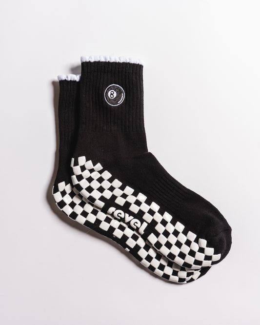 A pair of The Racked & Ready Grip Socks by Revel Athletic in black, featuring white ruffled cuffs, an 8-ball patch near the top, and a black-and-white checkerboard pattern on the soles. The word "Level" is printed on the bottom of these Pilates grip socks, combining stability and style for your workouts.