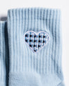 A close-up of The Cross My Heart Grip Sock by Revel Athletic in light blue, showcasing a ribbed design and an embroidered heart patch with a navy blue and light blue checkered pattern inside.